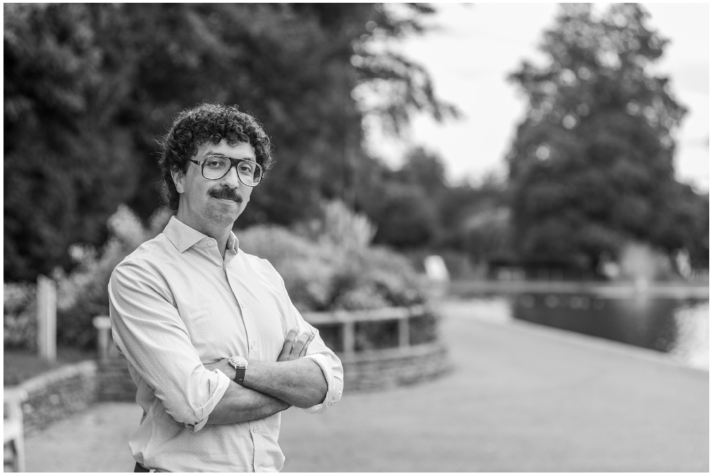 Black and white environmental portrait taken beside a waterside path in a park setting. The subject stands confidently with folded arms while the blurred trees, water and landscaped gardens provide context and depth. The monochrome treatment emphasises texture, expression and composition, with strong subject separation created by a shallow depth of field.